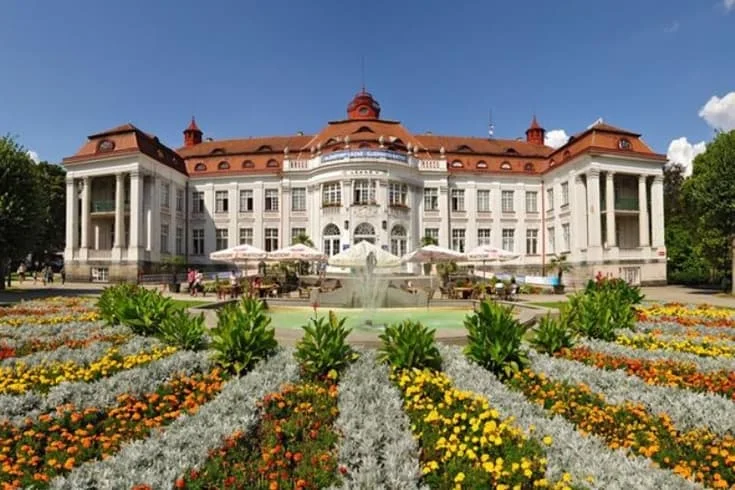 Velký lázeňský dům s fontánou před ním na pozadí barevných květin.