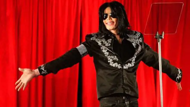 Michael Jackson v černém obleku na pódiu před červenou záclonou.