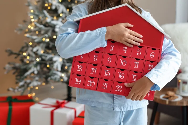 Dětské ruce drží červený adventní kalendář v podobě domečku.
