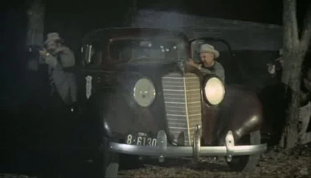 Stará policajní auta s ozbrojenými muži v kloboucích z filmu o Bonnie a Clyde.