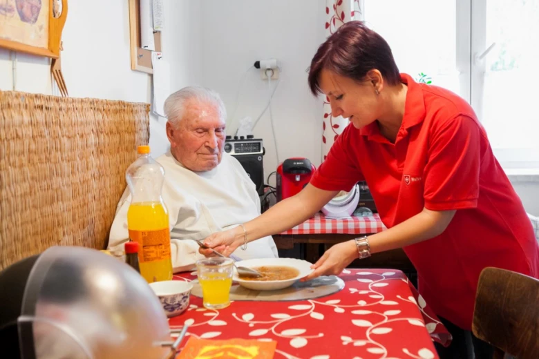 Péče o seniory: pracovnice podává jídlo seniorovi.