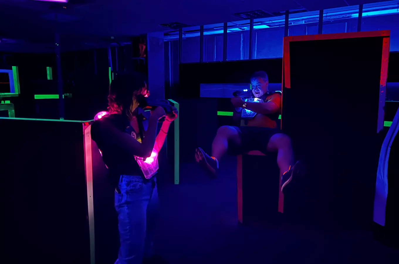 Dva lidé hrají vnitřní lasertag ve tmavém, neonově osvětleném prostoru.