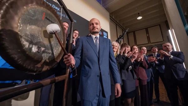 Muž v obleku buší do velkého zvonu před skupinou lidí.