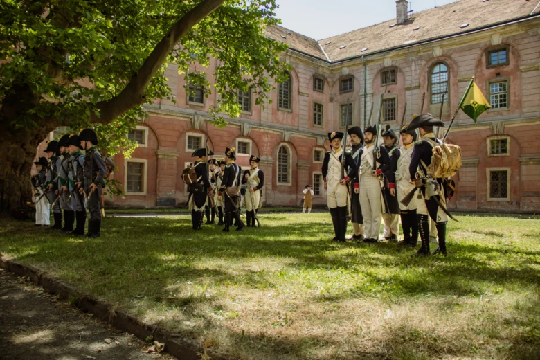 Skupina lidí v historických uniformách na trávníku před budovami.