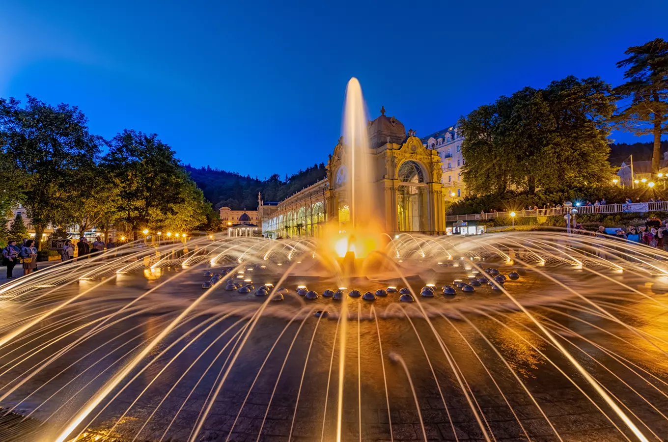 Zpívající fontána v Mariánských lázních za soumraku s osvětlenými vodními tryskami.