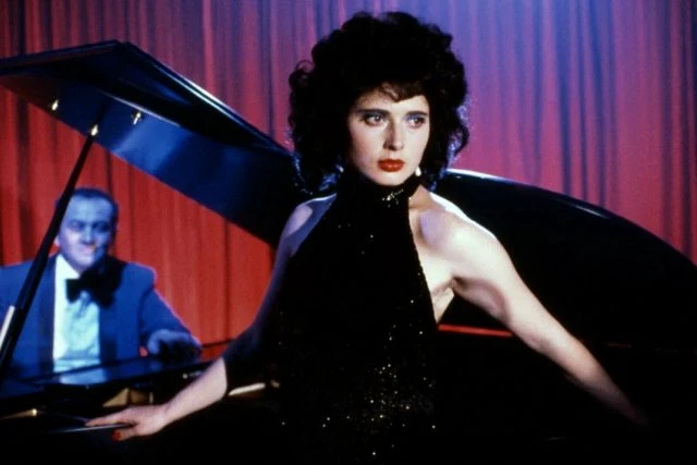 Isabella Rossellini v černém šatě u klavíru s mužem na pozadí.