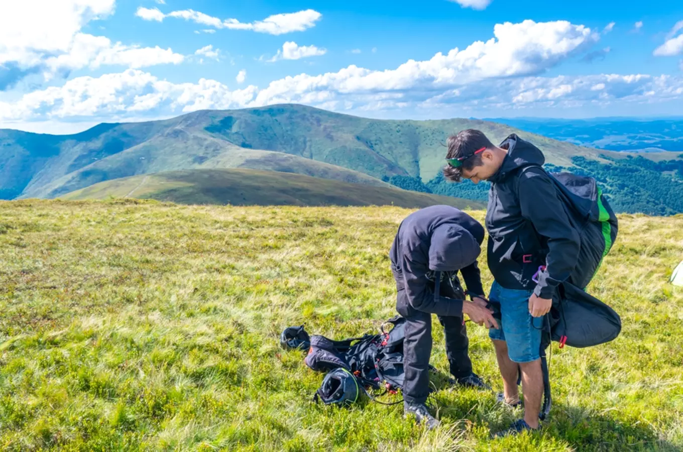Dva lidé připravují výstroj pro paraglidování na travnatém svahu.