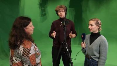 Tři moderátoři v televizním studiu s mikrofony před zeleným pozadím.