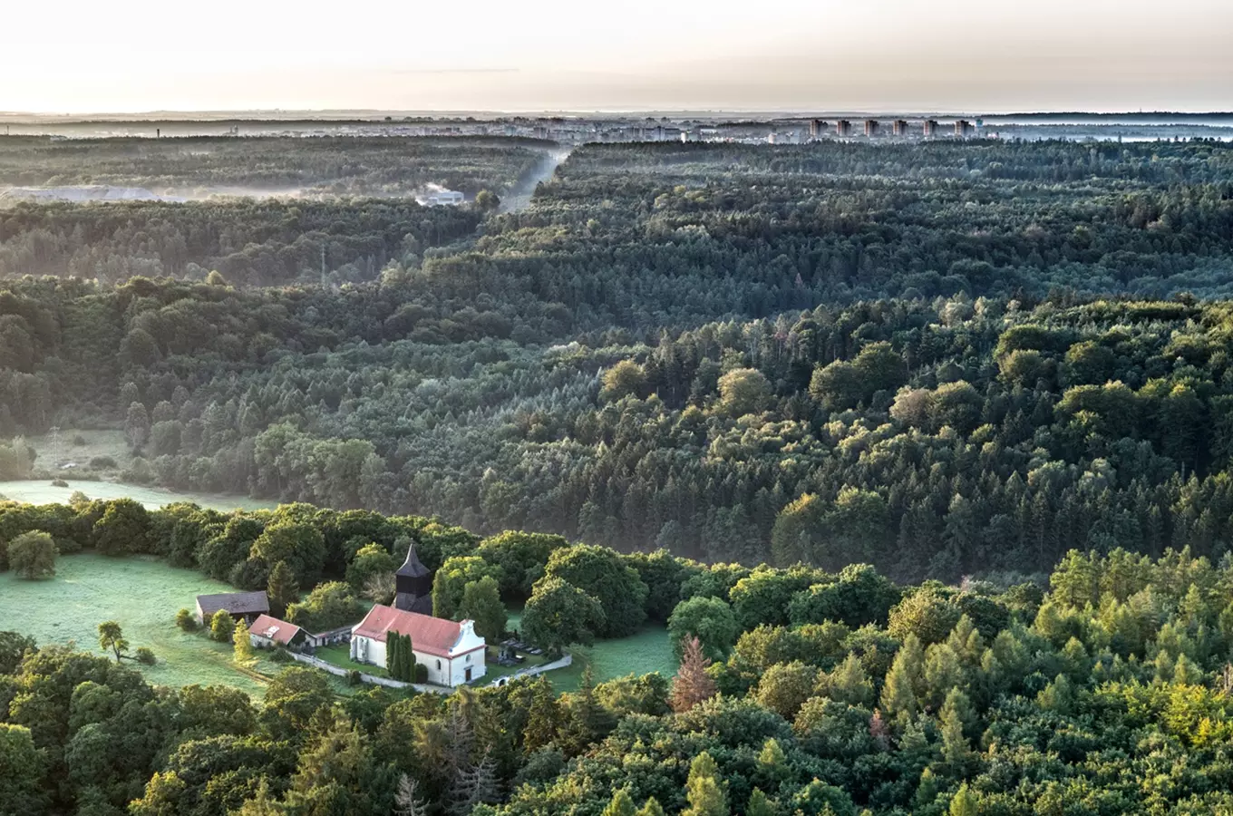 Lesní krajina s kostelem v popředí a Prahou na obzoru.