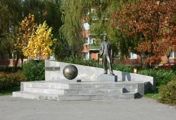 Bronzová socha muže s kulou na kamenném podstavci v parku.