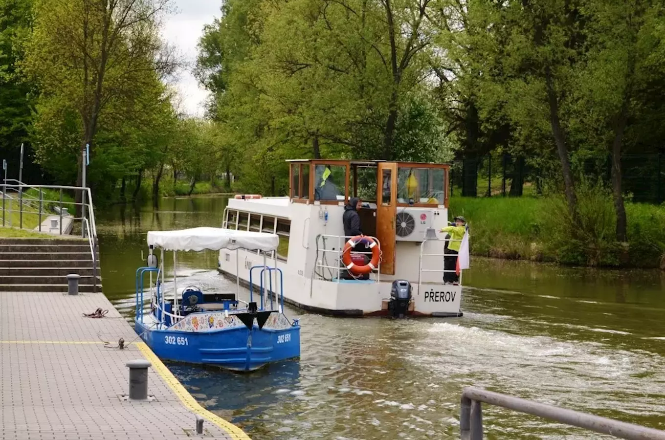 Bílá turistická loď a modrá pedální člun u mola na řece.