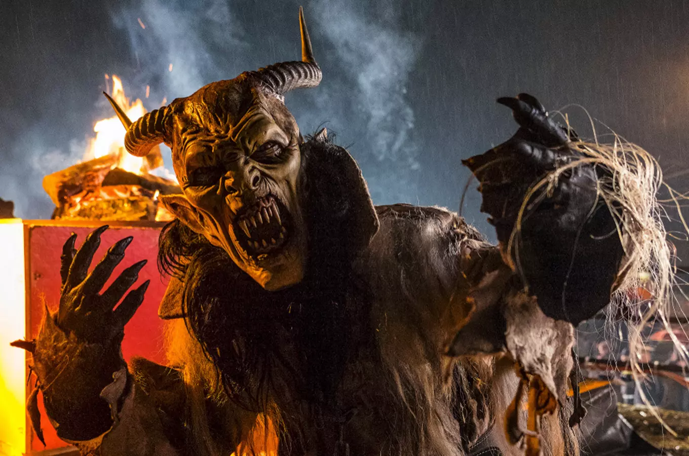 Krampus v kostýmu s rohy a vlasy na pozadí ohně.
