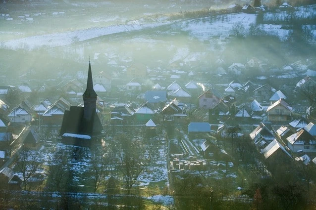 Sněhová rumunská vesnice s dřevěnou kostelem v mlze.
