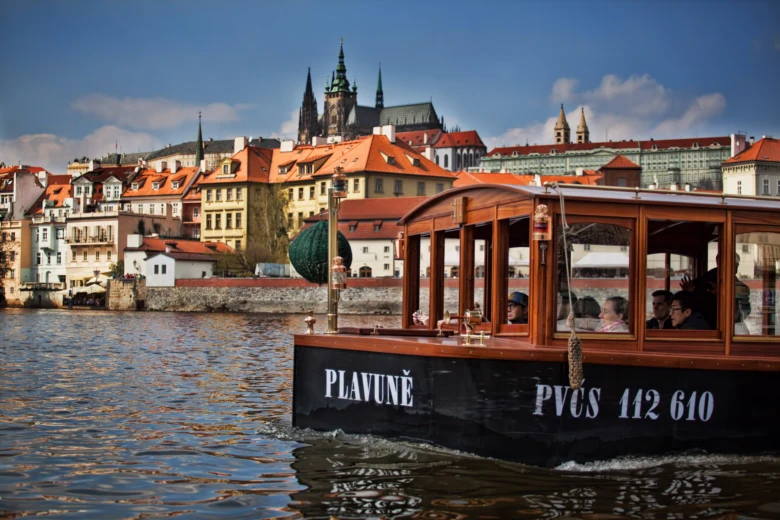 Turistická loď pluje po řece s historickým městem Prahou na pozadí.
