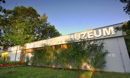 Bílá budova muzea s názvem MUZEUM obklopená zelenými stromy.