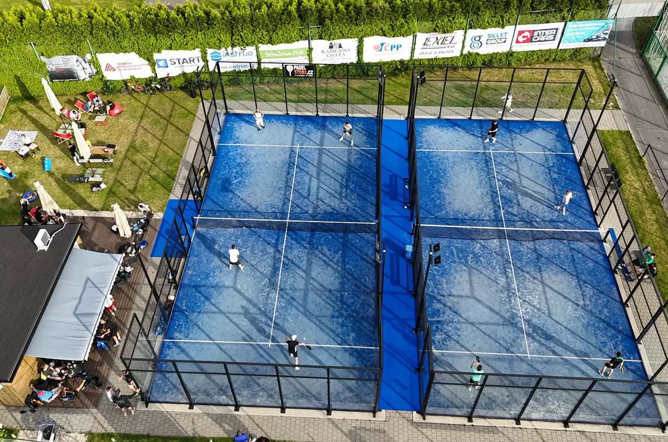 Dva modré dvorce na padelu s hráči a diváky venku.