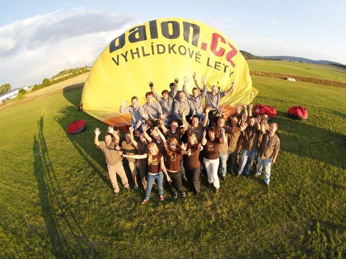 Skupina lidí stojí před nafukovaným žlutým balónem na louce.