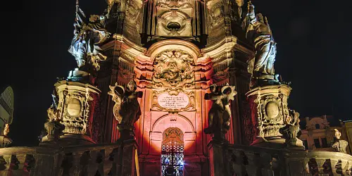 Noční pohled na osvětlený barokní pomník s sochami v Olomouci.