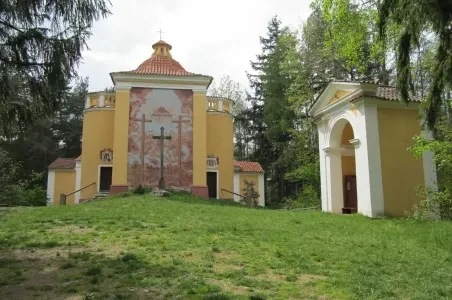 Žlutý kostel s kupolí vedle kapličky na trávníku mezi stromy