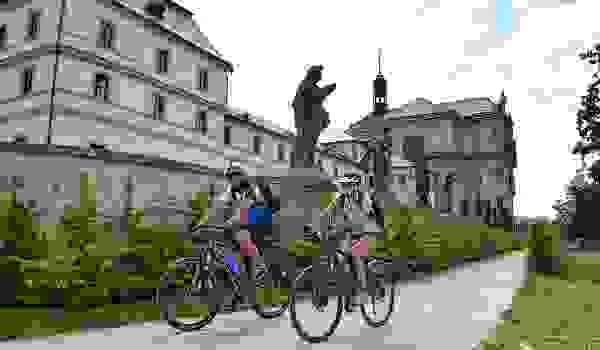 Cyklisté jedou po chodníku v Hradci Králové u sochy.