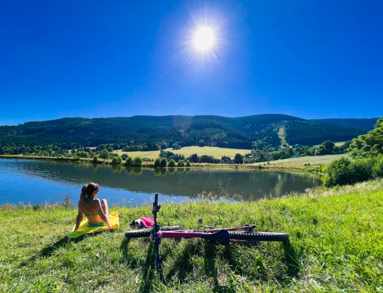 Žena sedí u jezera s horským bicyklem na pozadí hor.