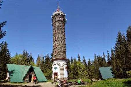 Kamenná věž v Jeseníkách s chatami uprostřed lesa.
