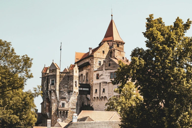 Historický český hrad s věžemi a červenými střechami mezi stromy.