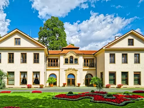 Velká historická budova s červenou střechou na zelené trávě.