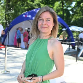 Žena v zeleném šatech stojí venku s brýlemi.