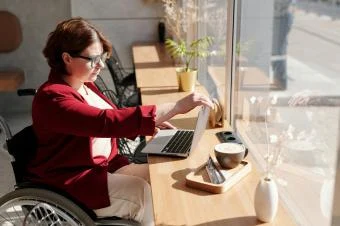 Žena v invalidním vozíku pracuje na počítači u okna.