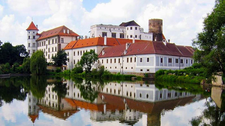 Bílý hrad s červenou střechou odražený v klidné vodě.
