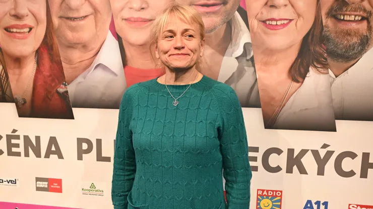 Žena v zeleném svetrku stojí před plakátem s českým nápisem.